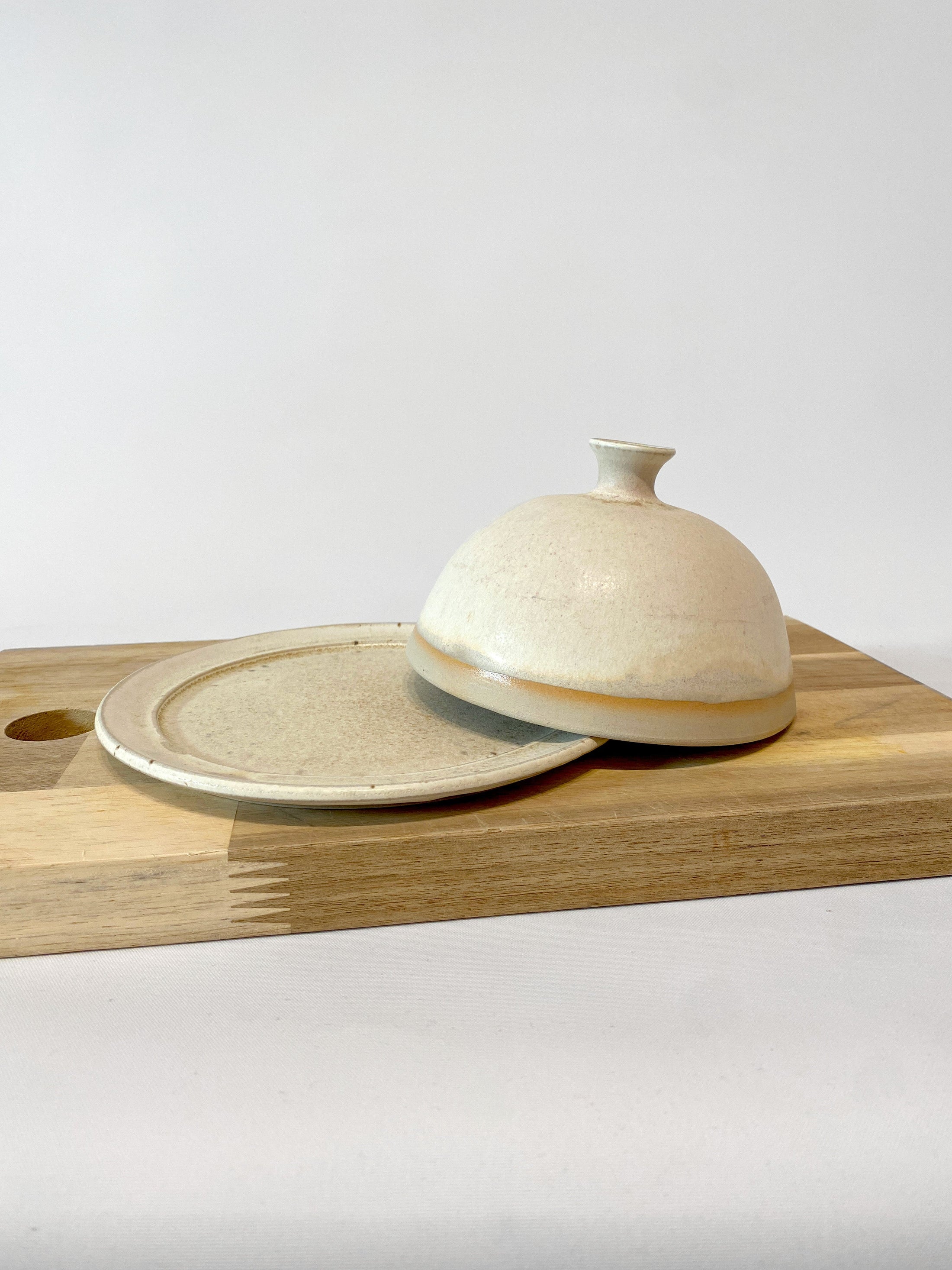 Geöffnete, runde Butterdose in Beige mit knopfförmigem Griff auf Holzbrett